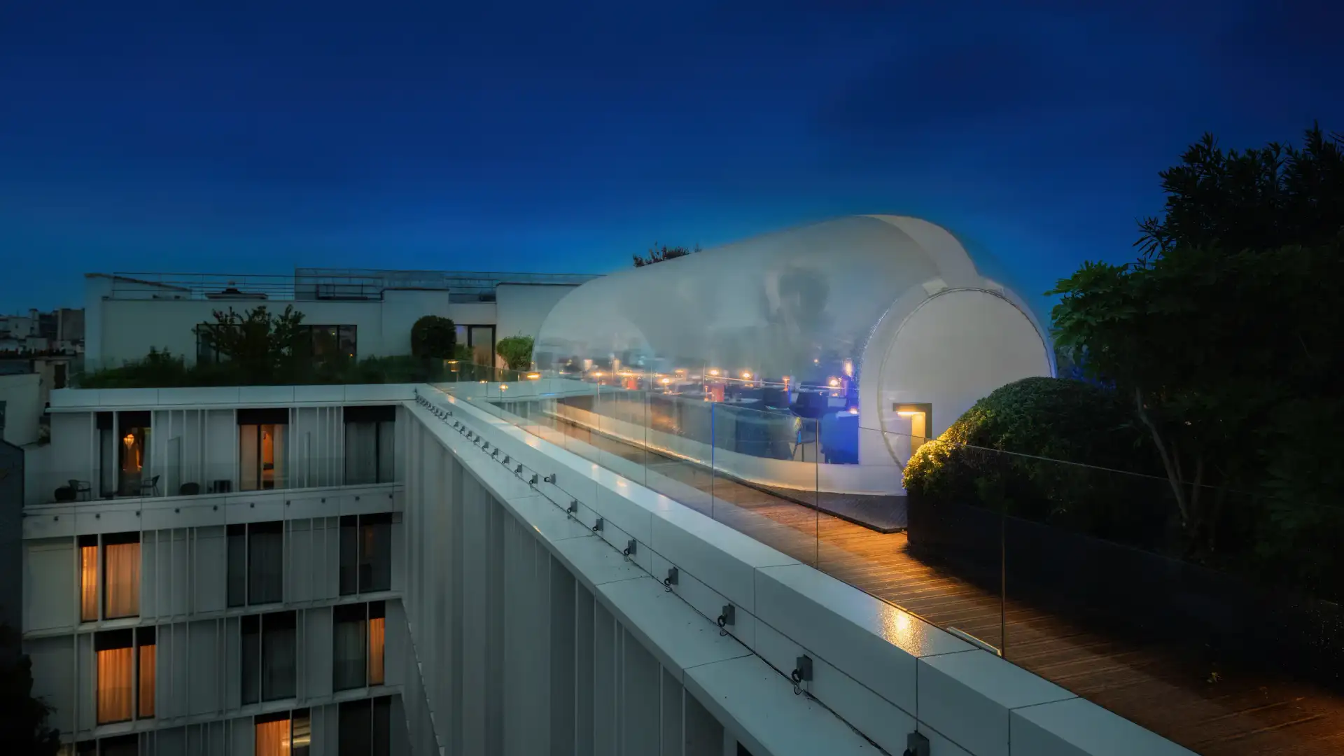 A wooden illuminated walkway leads to a translucent modern oval-shaped building, with residential buildings in the background.
