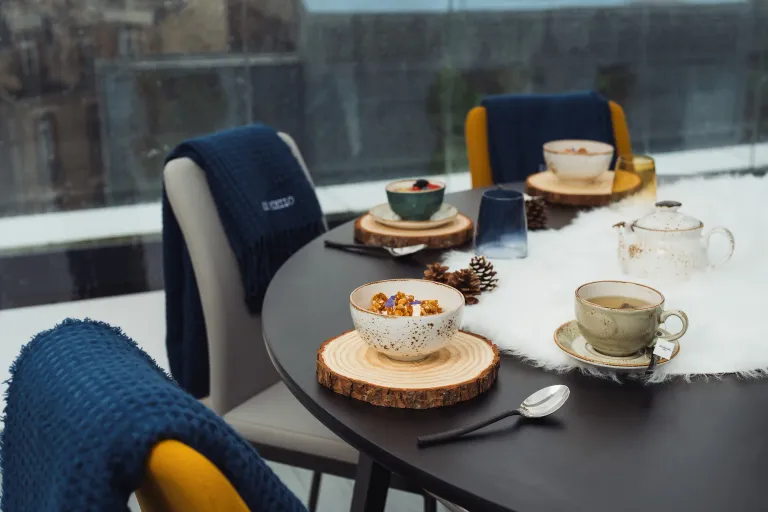 Table dressée avec des bols de nourriture, une théière et des tasses, sur une nappe blanche avec des éléments de décoration en bois et des coussins sur les chaises
